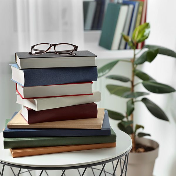 Stack of books with glasses on top, resting on a round table. A potted plant and bookshelf are in the background.